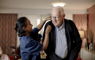 Senior man looking at female nurse helping him to wear sweater in retirement home